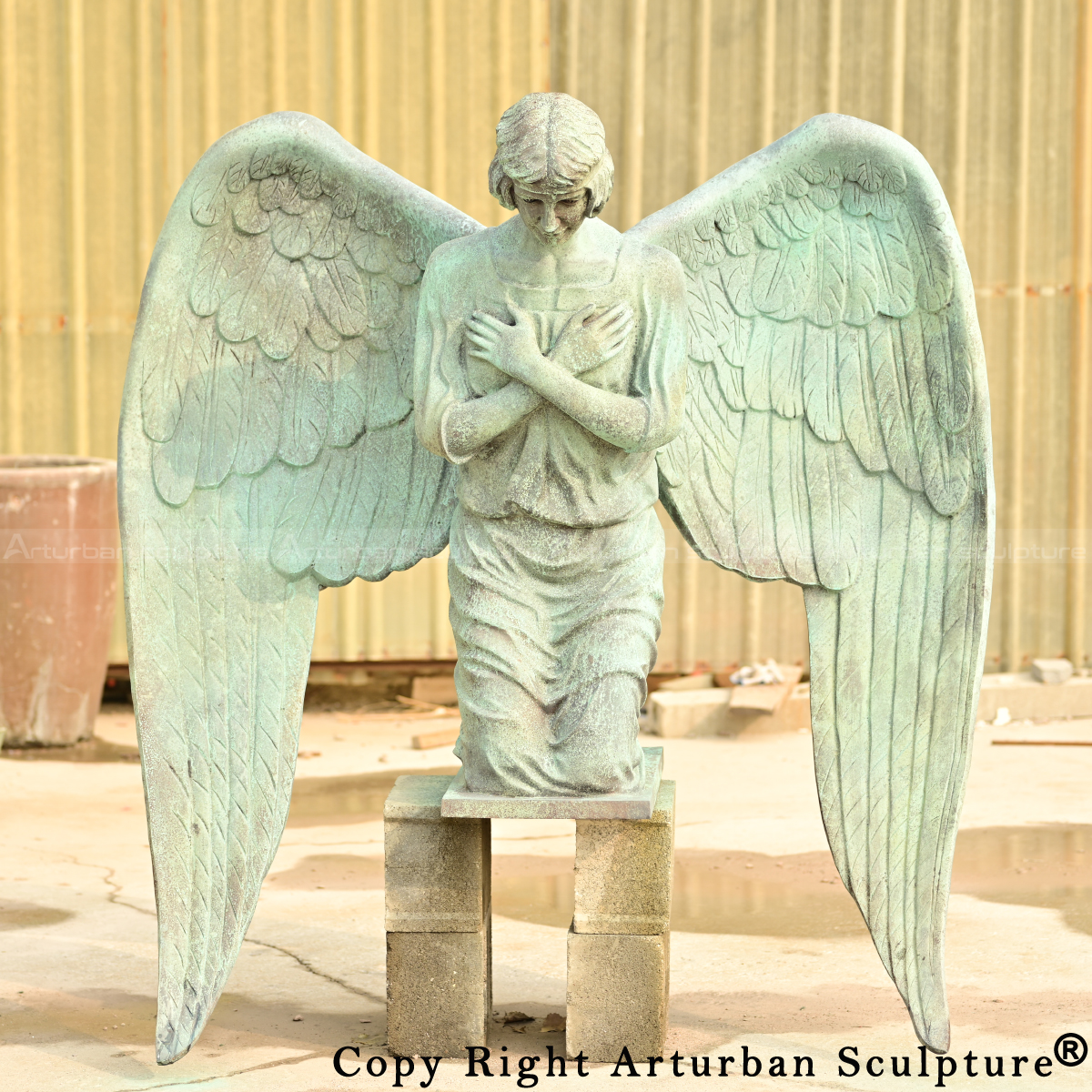 Frontal view of the bronze kneeling angel statue with folded arms and solemn expression, featuring a green patina finish