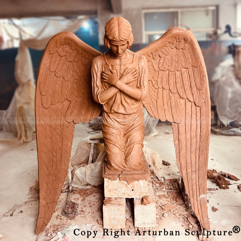 Frontal view of the clay model of a kneeling angel sculpture with folded arms and downward gaze