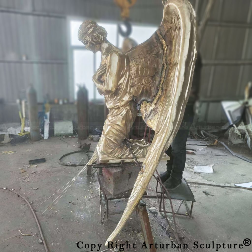Bronze kneeling angel statue during welding process, showing one side of the body with folded hands and extended wings