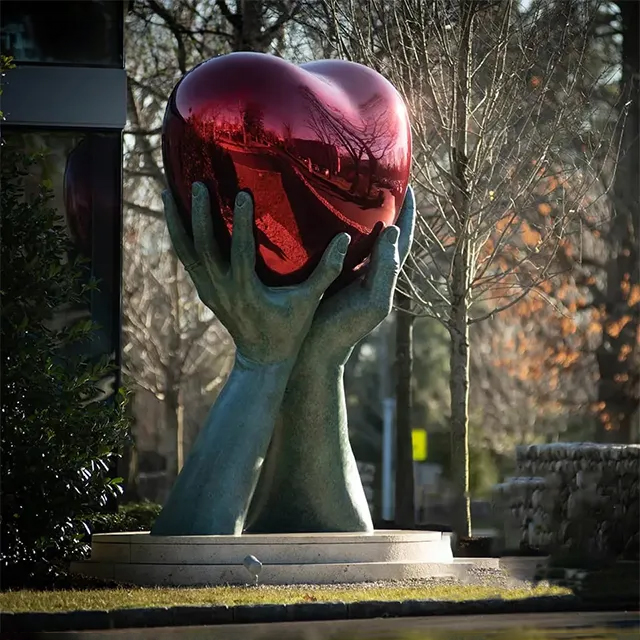 Hands holding a red Stainless Steel Heart Sculpture outdoors by Lorenzo Quinn